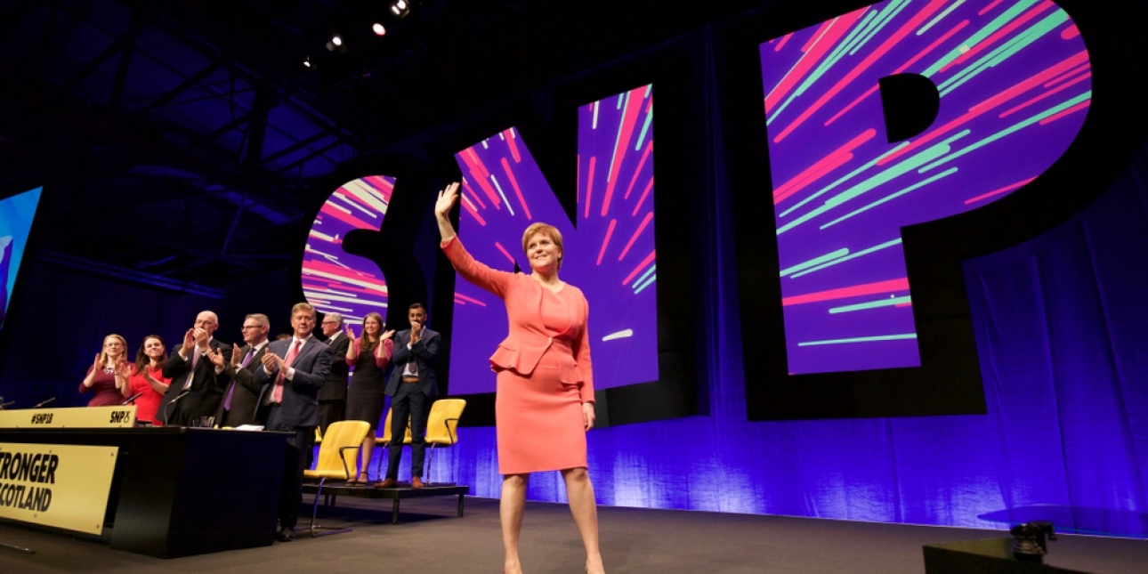 A smiling Nicola Sturgeon waving to the audience while on stage at a SNP conference. She is wearing a coral outfit. There is are large SNP letters behind her and a selection of delegates on stage giving a standing ovation